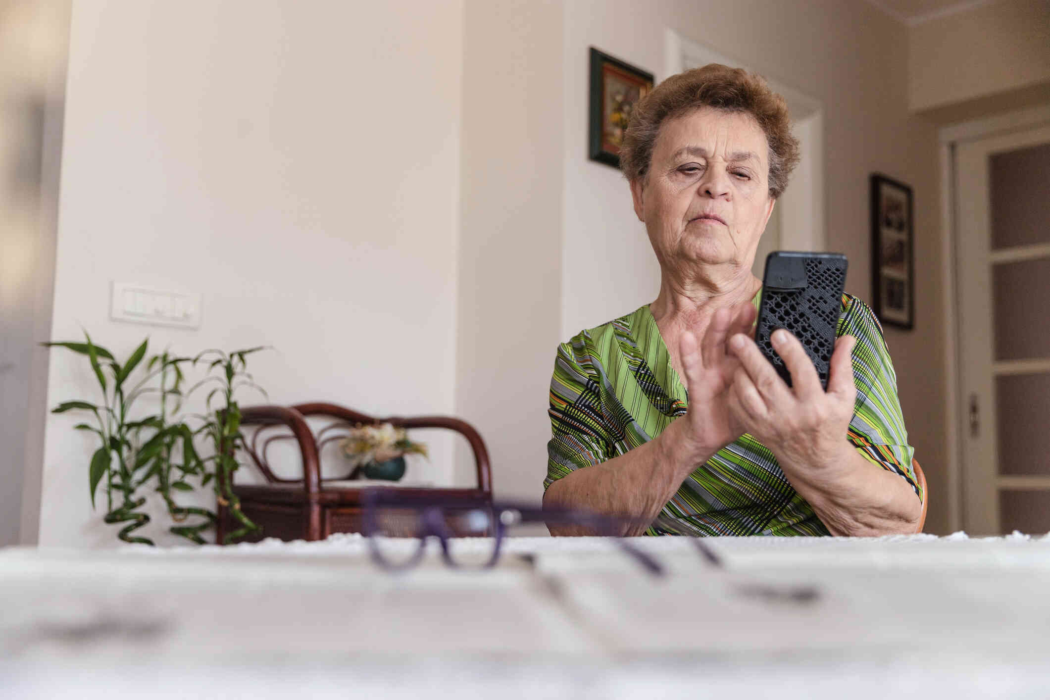 An elderly woman in a green shirt sits at the kitche table and taps on the cellphone in her hand with a focused expression.